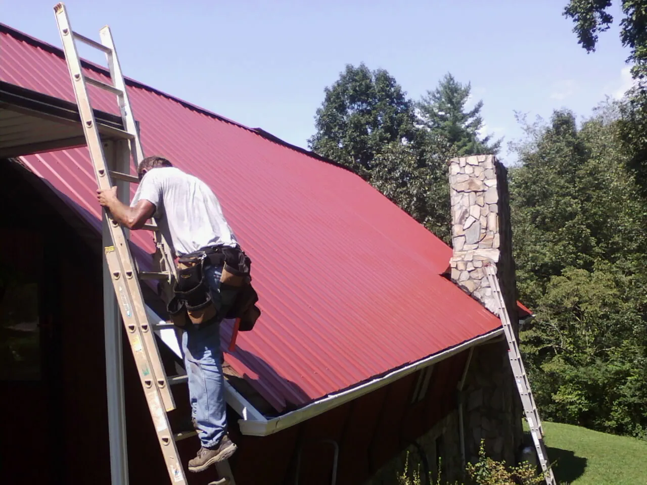 Roofer on a ladder beside a red metal roof and stone chimney — Soffit & Fascia Repair by Roofing Brothers in Jefferson